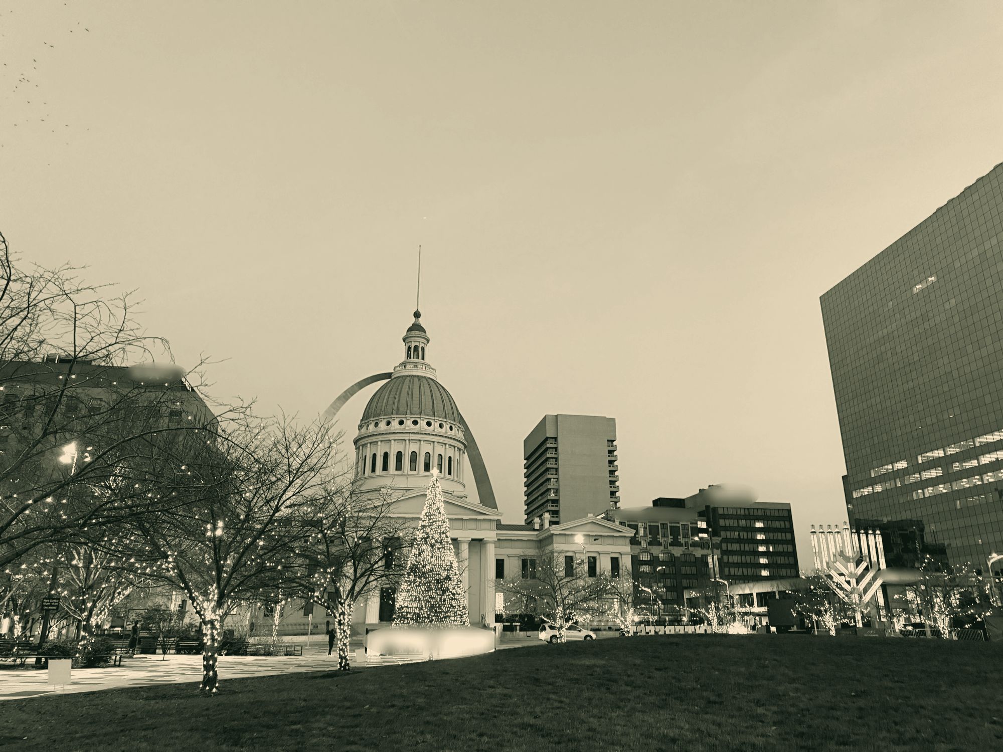 The Old Courthouse dome framed by the Gateway Arch at dusk, with a lit holiday tree and string-lit trees in the park.