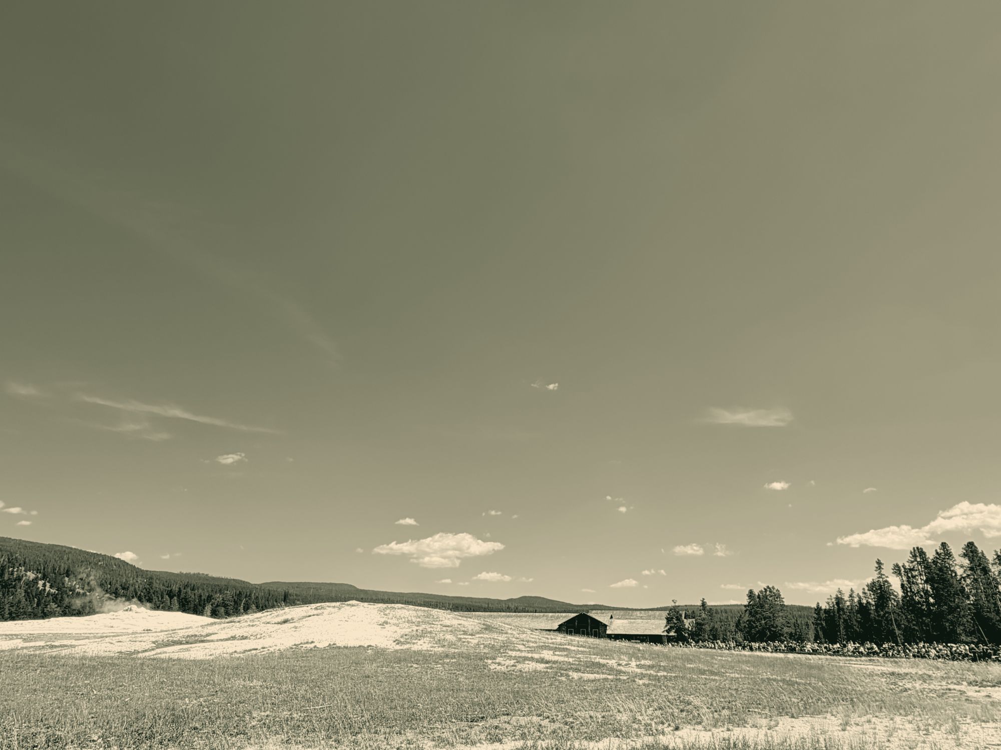 A log lodge at the edge of a wide geyser basin under a huge sky, steam rising at the left.