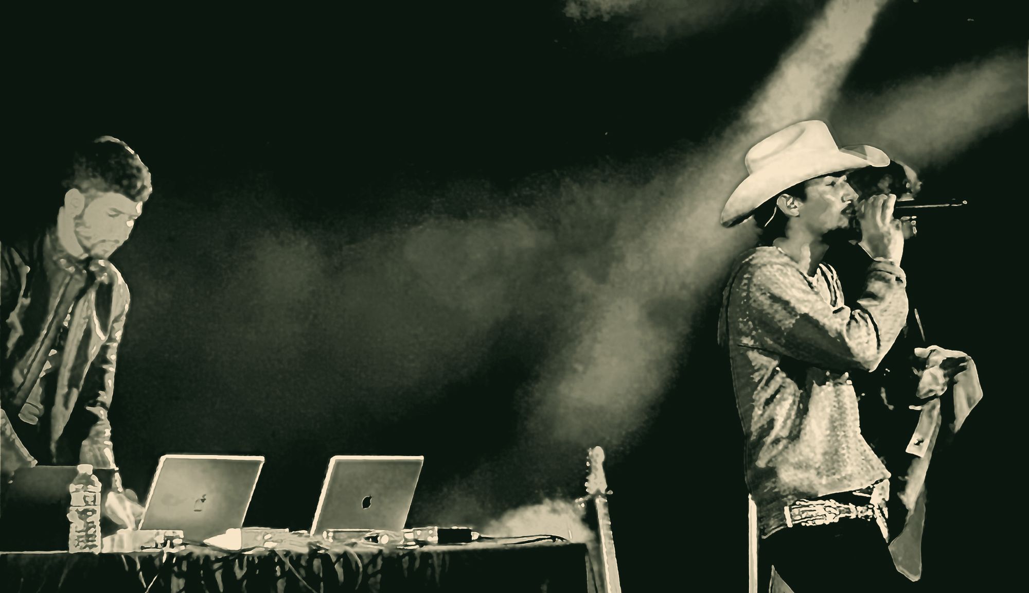 Two performers on a dim stage in black and white: one working at two laptops, the other in a cowboy hat singing into a microphone through stage smoke.